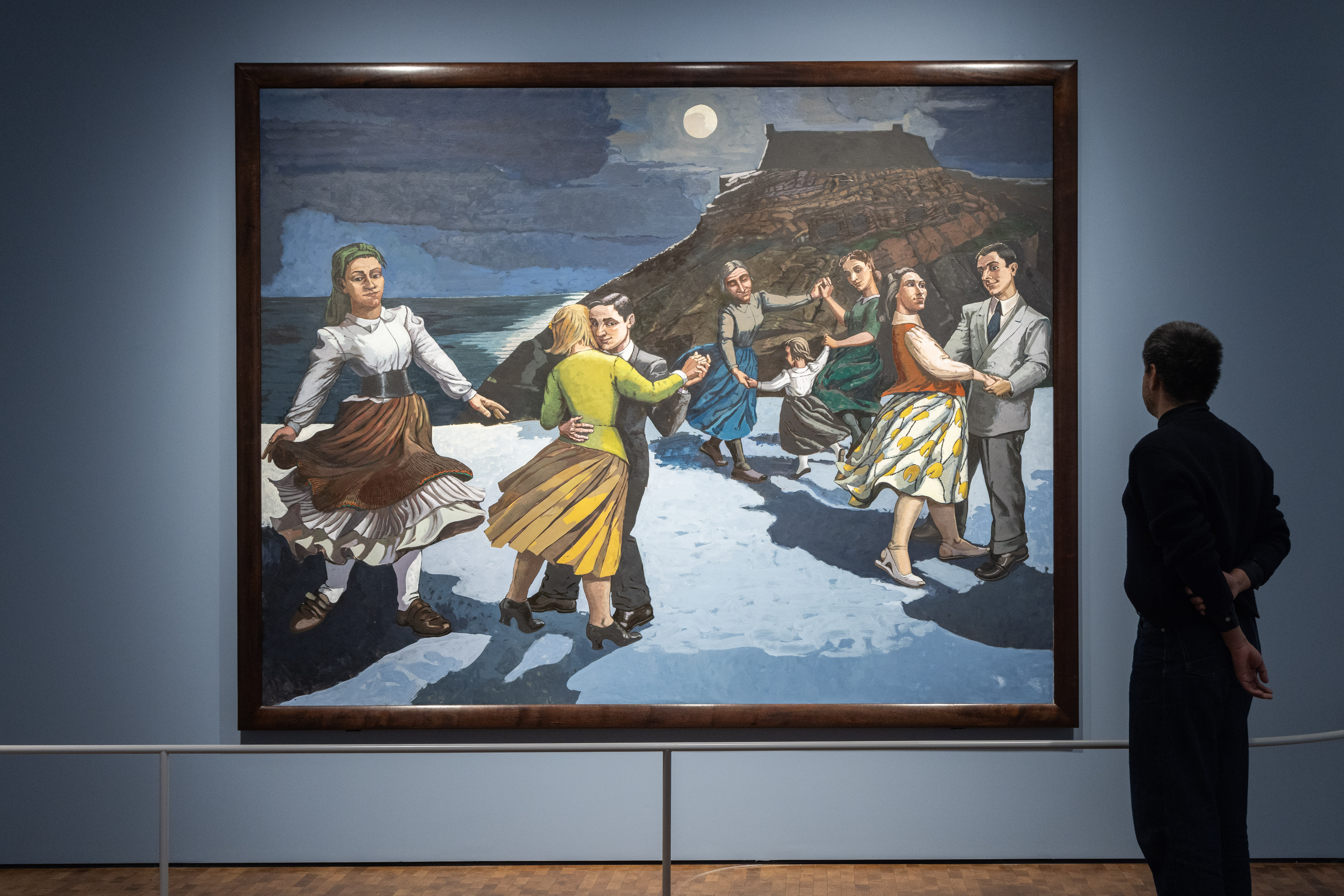 En mann står med ryggen mot kamera og betrakter verket Dansen av Paula Rego. Maleriet viser en strand med et fjell i bakgrunnen og en mørk himmel med en lysende måne. På stranden er det flere par som danser og en kvinne som danser alene. Fotografi