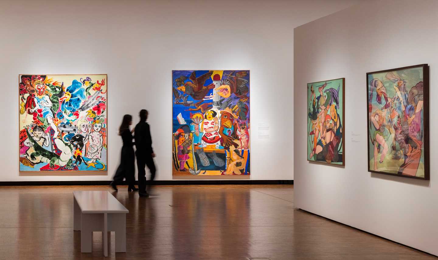 Bilde fra utstillingssal som viser verk av Paula Rego. I dette bildet er det fire fargerike verk som henger på hvite vegger. To mennesker går gjennom salen. Fotografi