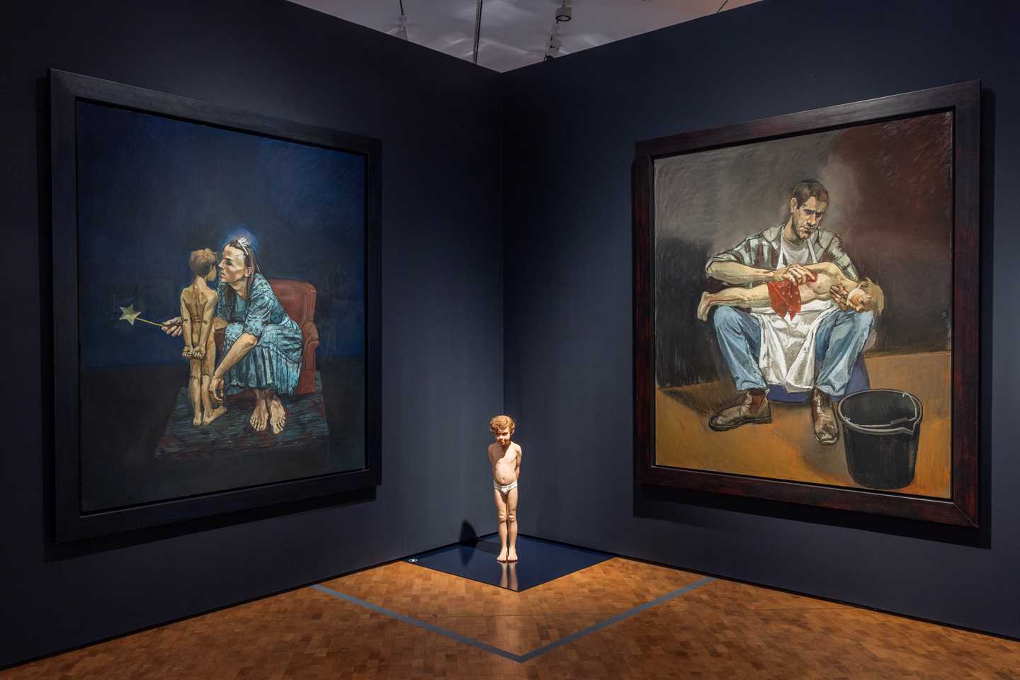Mot en mørkeblå vegg henger to store verk der det ene viser en kvinne sammen med en liten gutt, og det andre en mann sammen med den samme gutten. På gulvet midt mellom pastellene står en skulptur av den lille gutten. Fotografi