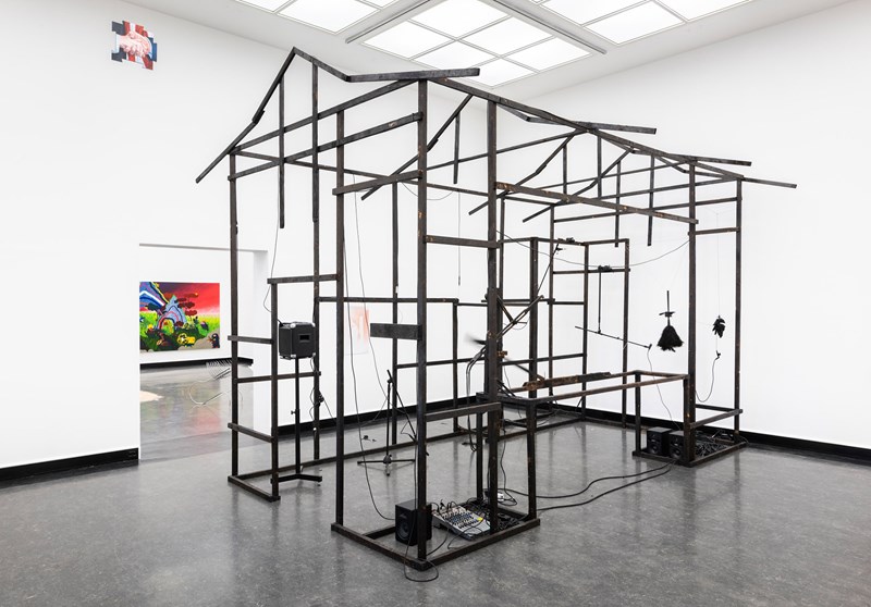 In a white gallery space, many black rods form the framework of a small, elongated house. Photograph.