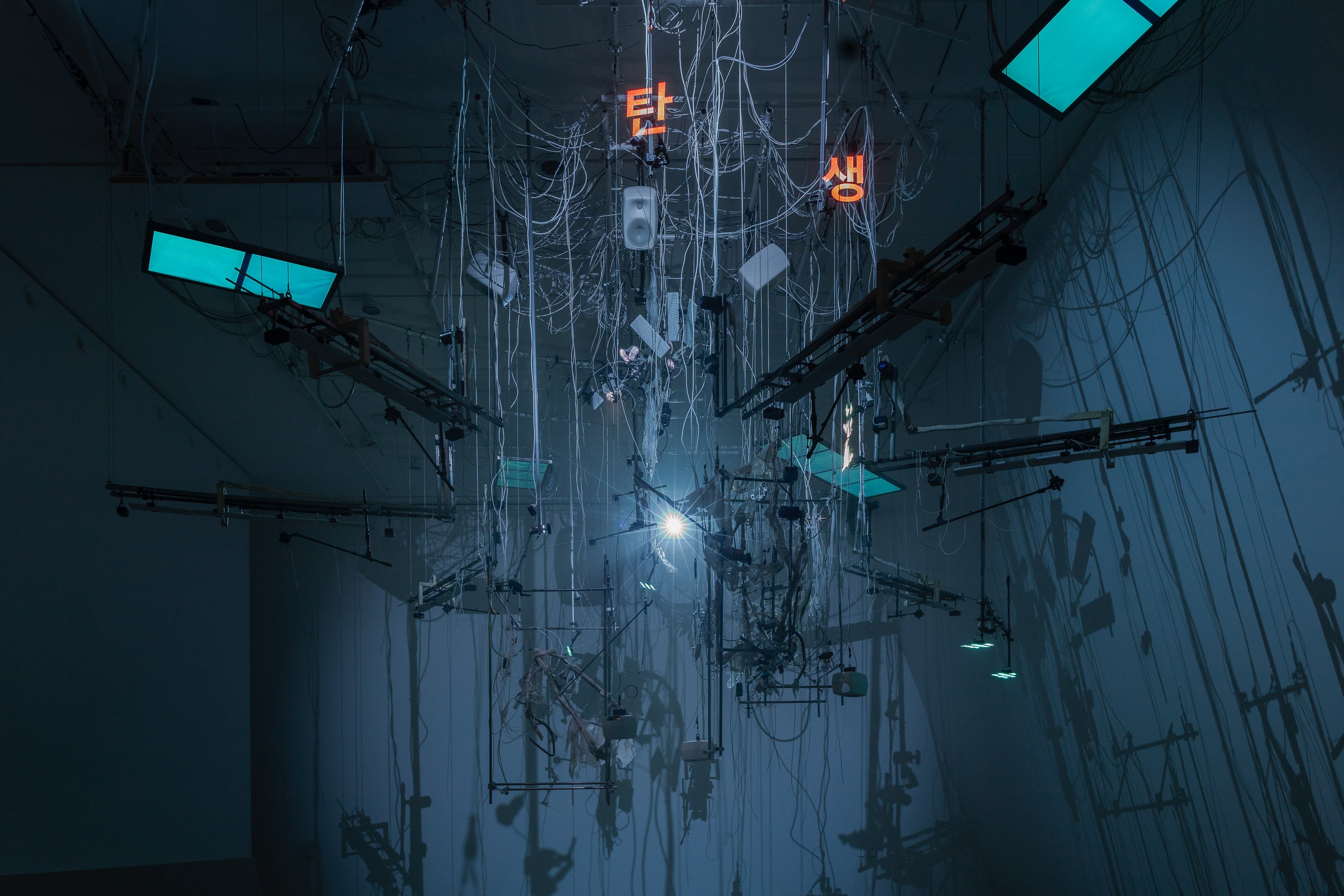 En installasjon henger ned fra taket i et mørkt rom lyst opp av blått lys. Installasjonen består blant annet av mange ledninger, strenger til musikkinstrumenter, lysskjermer og tastaturer. Fotografi