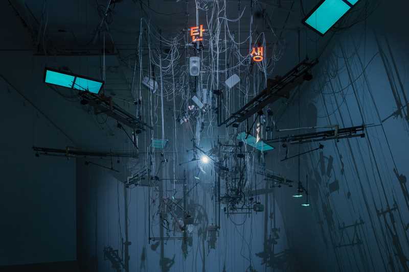 An installation hangs from the ceiling in a dark room illuminated by blue light. The installation includes numerous wires, instrument strings, light screens, and keyboards. Photograph.