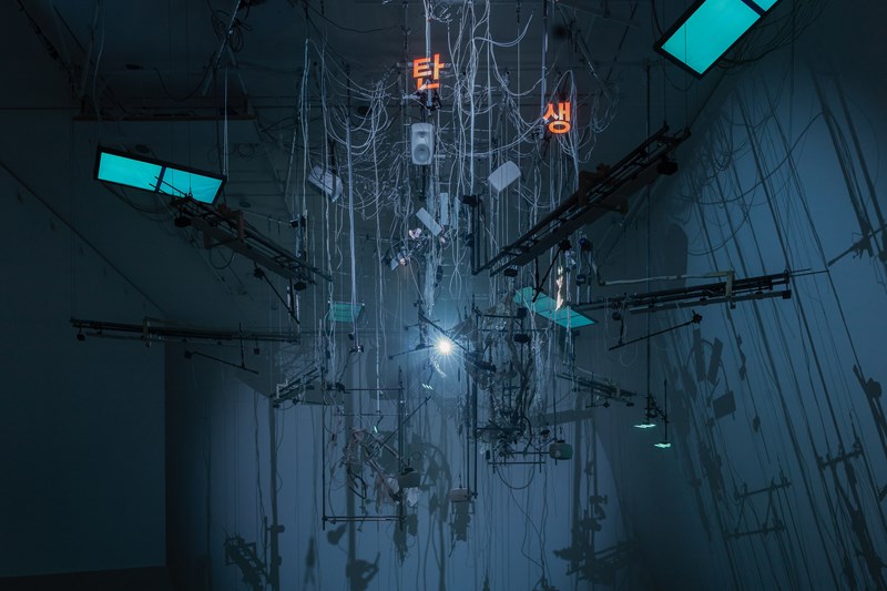 En installasjon henger ned fra taket i et mørkt rom lyst opp av blått lys. Installasjonen består blant annet av mange ledninger, strenger til musikkinstrumenter, lysskjermer og tastaturer. Fotografi