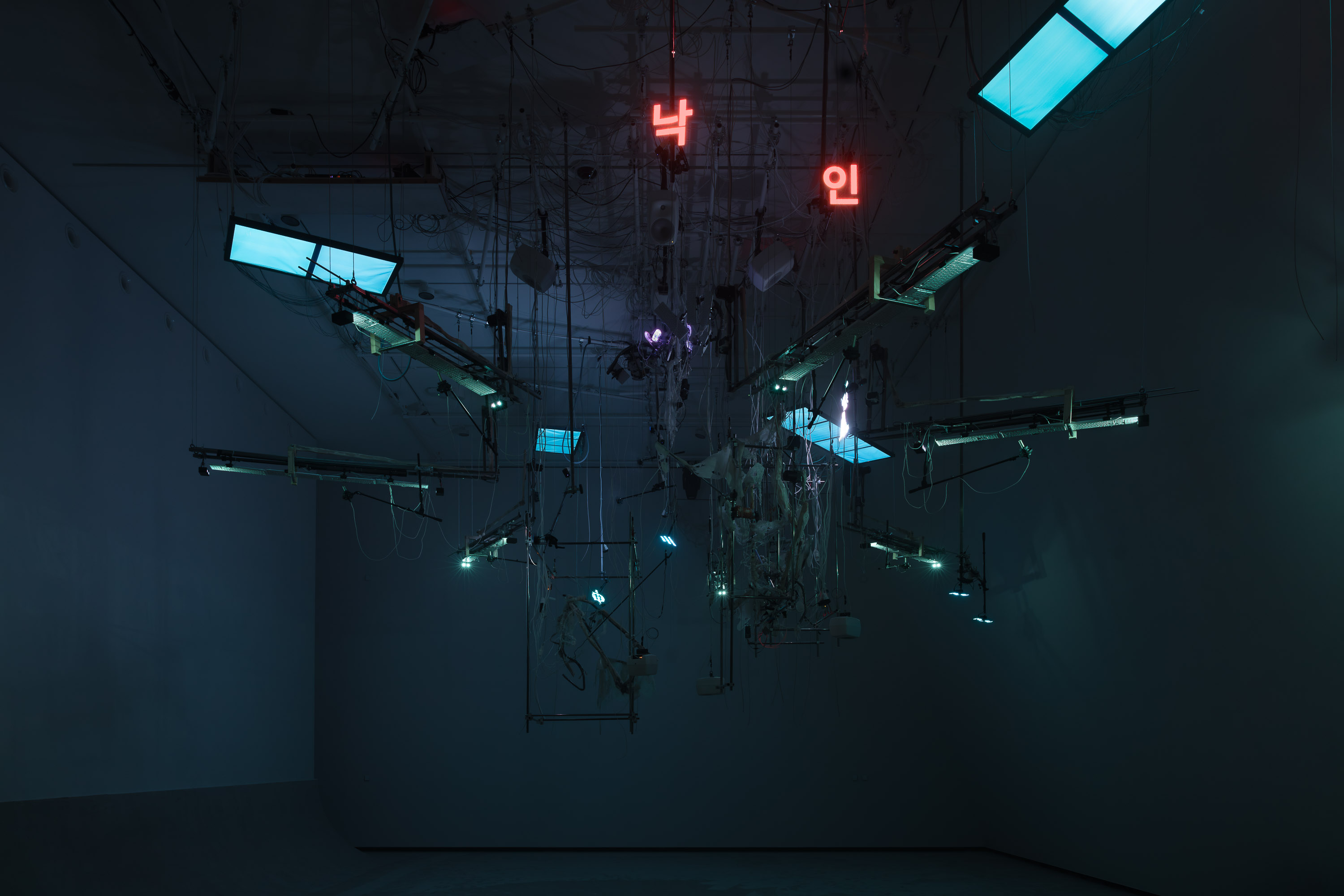 An installation hangs from the ceiling in a dark room illuminated by blue light. The installation includes numerous wires, instrument strings, light screens, and keyboards. Photograph.