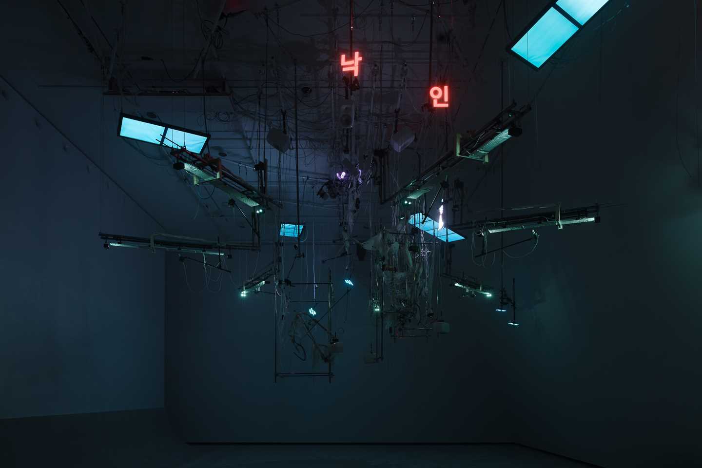 An installation hangs from the ceiling in a dark room illuminated by blue light. The installation includes numerous wires, instrument strings, light screens, and keyboards. Photograph.