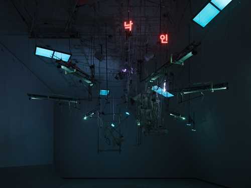 An installation hangs from the ceiling in a dark room illuminated by blue light. The installation includes numerous wires, instrument strings, light screens, and keyboards. Photograph.