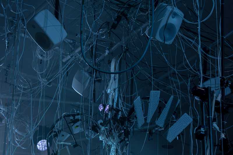 Speakers and keyboards hang from the ceiling in a tangle of wires and strings. Photograph.