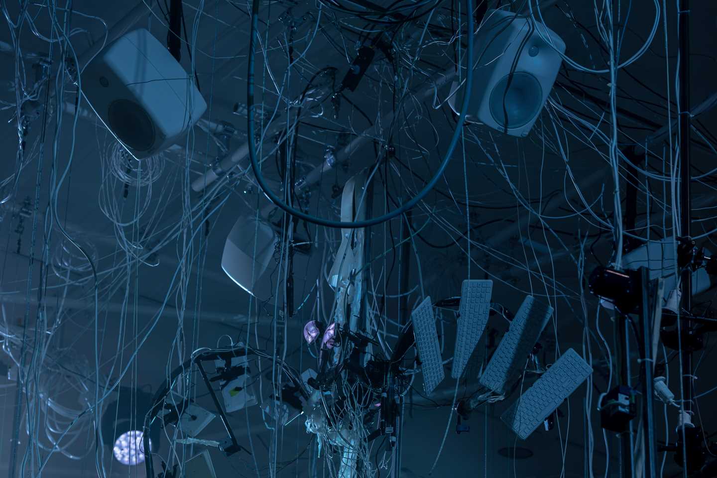 Speakers and keyboards hang from the ceiling in a tangle of wires and strings. Photograph.