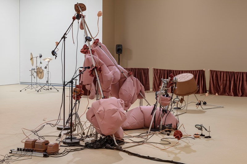 An installation in a room with sand-coloured and white walls. In the background, stands with cymbals are visible. In the foreground, stands fitted with pink fabric cushions can be seen. Wires run throughout, connecting different parts of the installation. Photograph.