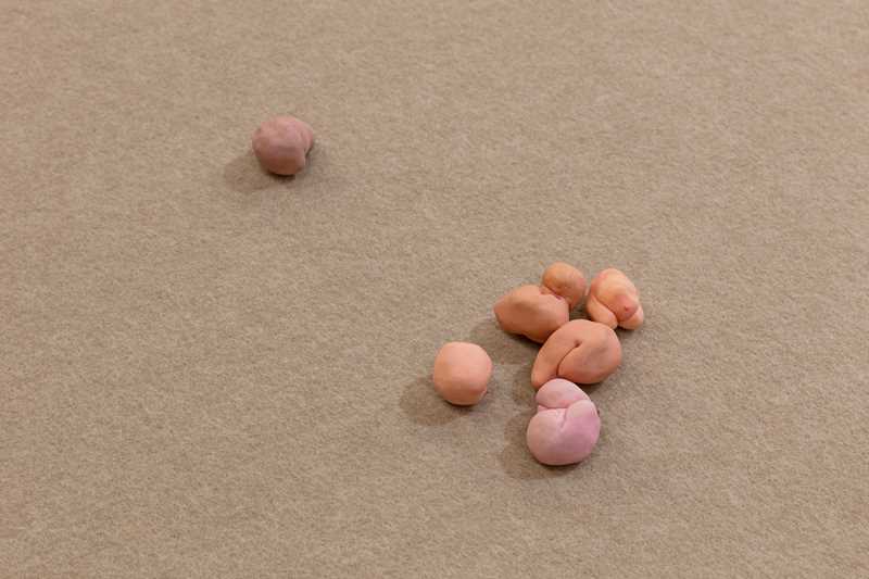 Close-up of part of the installation. On a sand-coloured floor lie small figures in shades of pink, orange, and brown that appear bodily in form. Photograph.