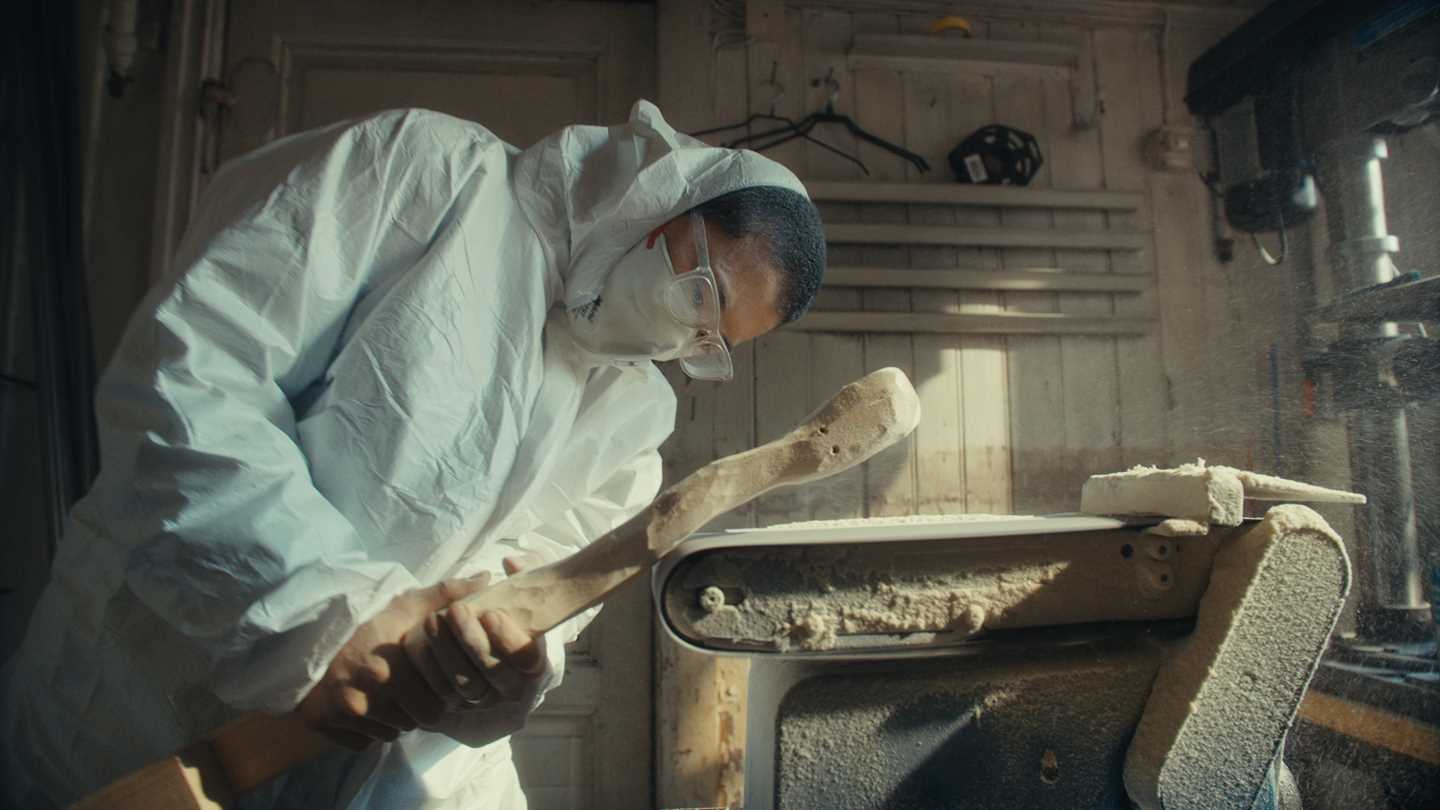 Kunstner Kim Hankyul i heldekkende hvit maledress, med munnbind og vernebriller står krumbøyd og sliper en trestokk på en automatisk slipemaskin. Fotografi