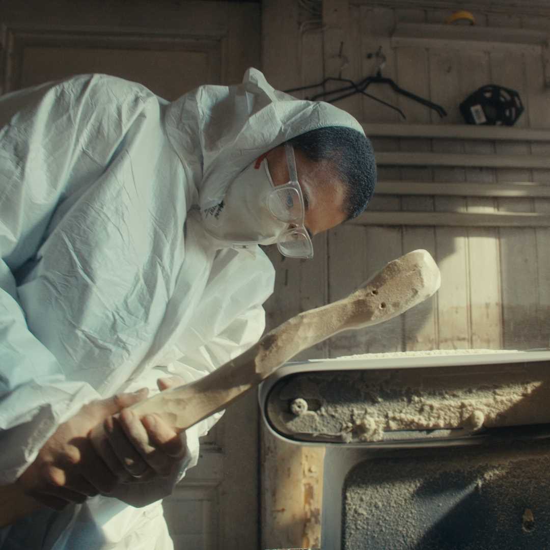 Kunstner Kim Hankyul i heldekkende hvit maledress, med munnbind og vernebriller står krumbøyd og sliper en trestokk på en automatisk slipemaskin. Fotografi