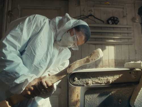 Artist Kim Hankyul in a full white coverall, mask and safety glasses, bent over sanding a log on an automatic sanding machine. Photograph.