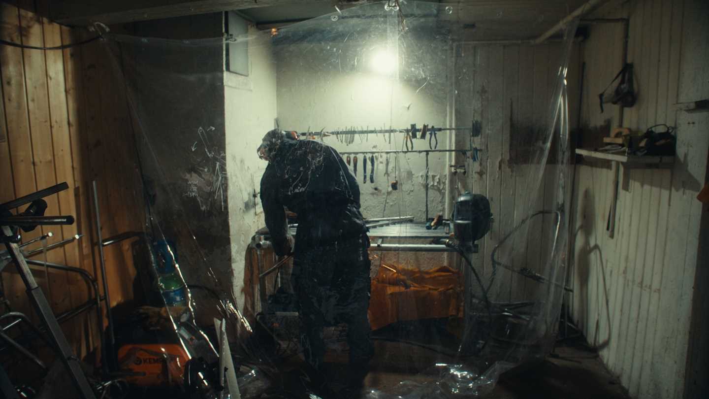 En mann står med ryggen mot kamera i et verksted. Han har på seg en arbeidsdress og det henger gjennomsiktig plast ned fra taket. Fotografi
