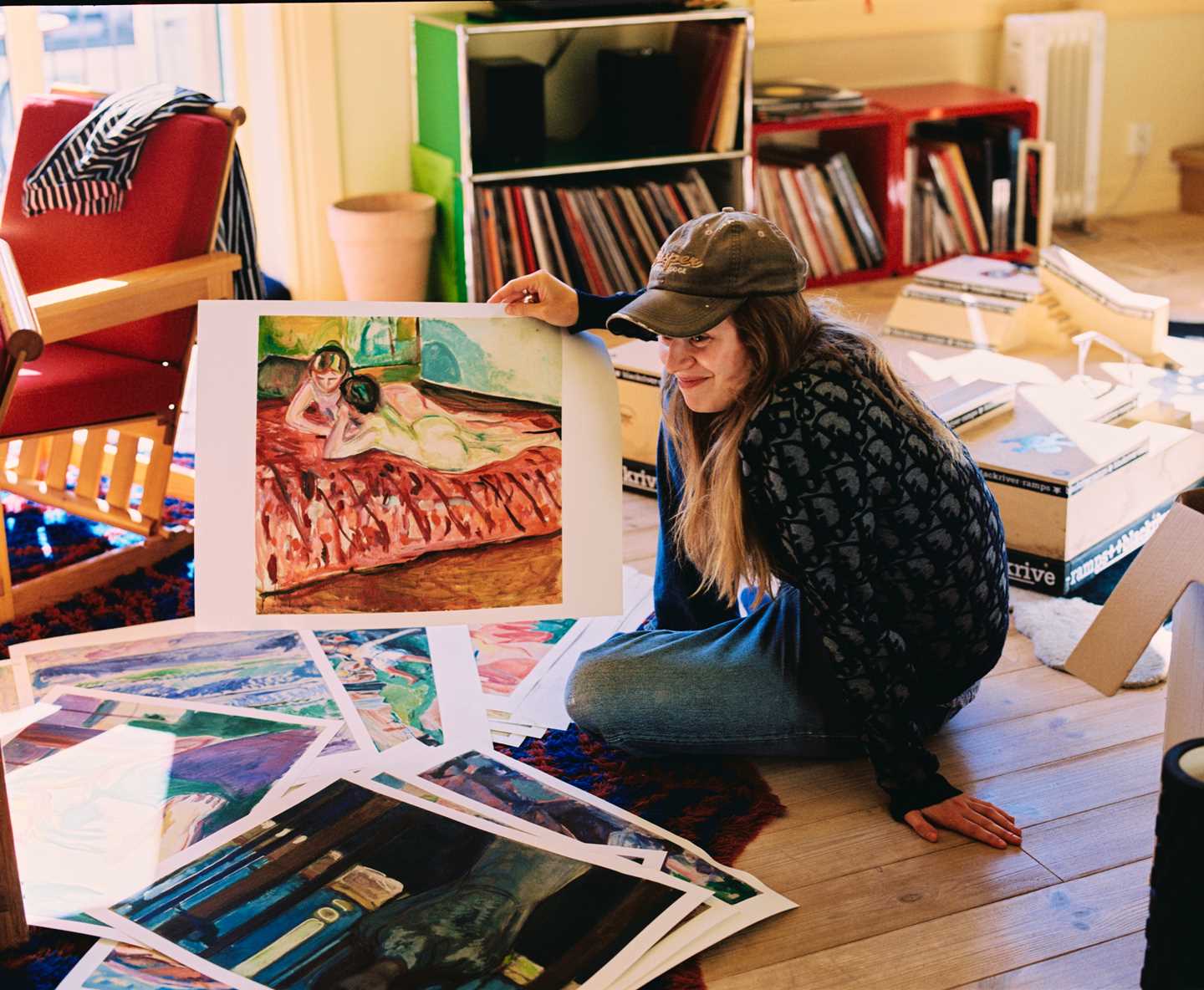 The artist Marie Ulven is sitting on the floor of a flat, surrounded by numerous posters featuring works by Edvard Munch. Marie is wearing a cap and holding up an artwork depicting two naked women on a bed. She appears to be on the verge of laughing. Photograph.