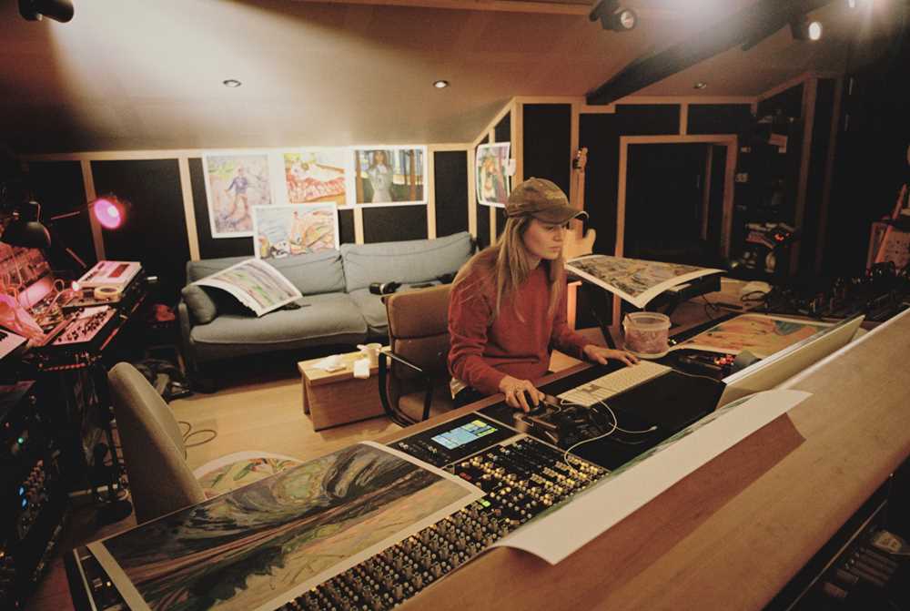 Artist Marie Ulven is seated at the mixing desk in her own studio. Posters featuring works by Edvard Munch are hanging on the wall behind her. Photograph.