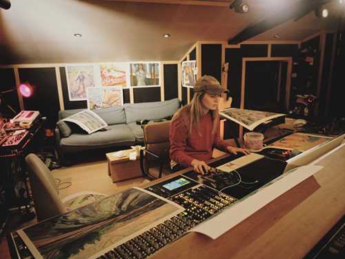 Artist Marie Ulven is seated at the mixing desk in her own studio. Posters featuring works by Edvard Munch are hanging on the wall behind her. Photograph.