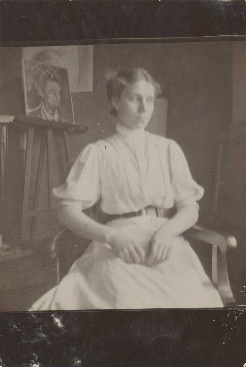 En kvinne i hvit kjole sitter med hendene i fanget og blikket vendt bort. I bakgrunnen vises Munchs atelier med et maleri på veggen og ett på staffeli. Fotografi