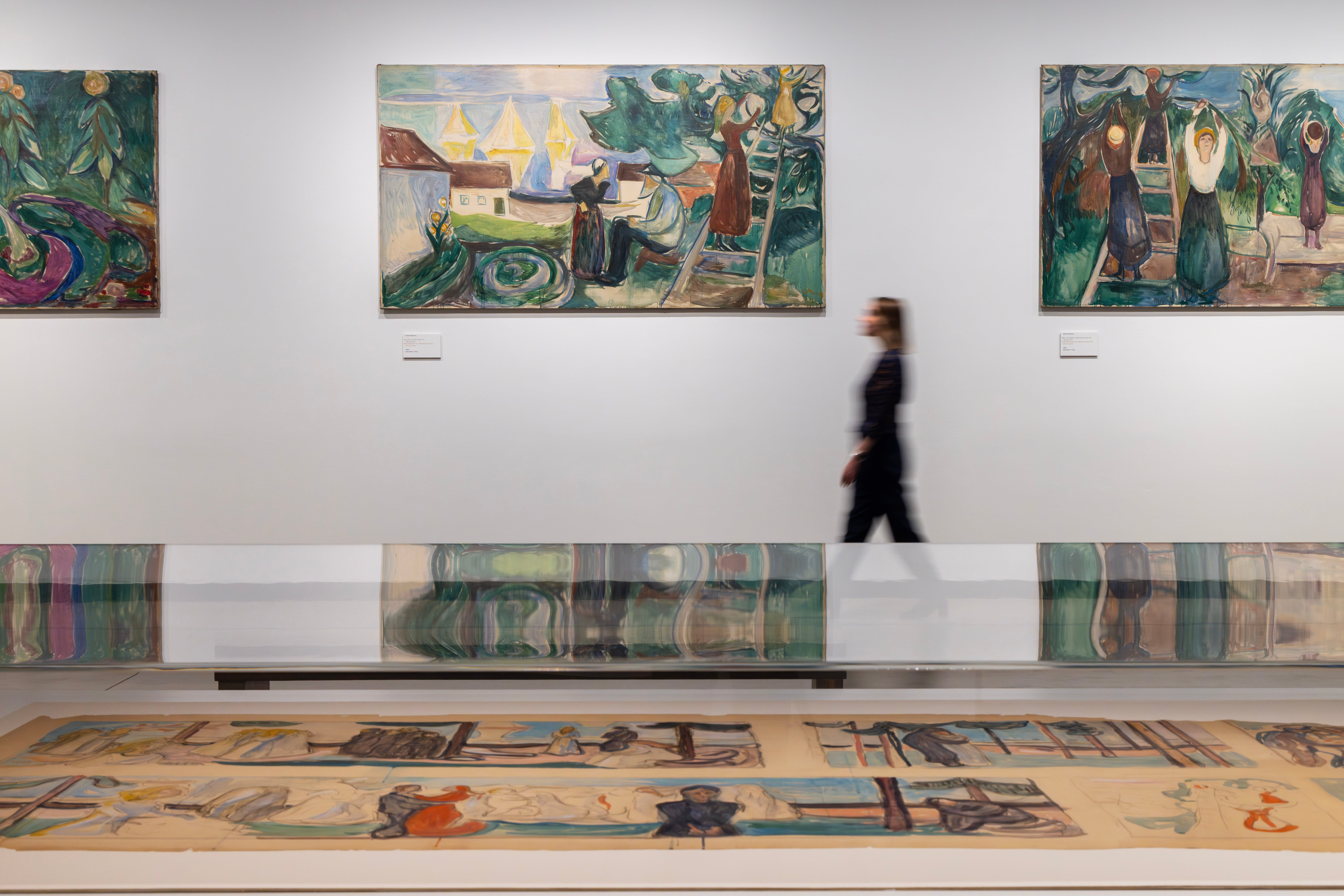 In the foreground is a glass display case containing sketches. In the background, a woman walks along a white wall while looking at the paintings hanging there. Photograph.