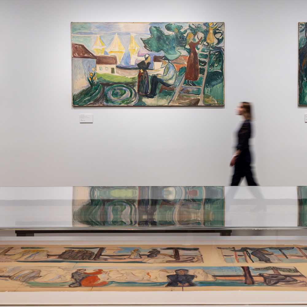 In the foreground is a glass display case containing sketches. In the background, a woman walks along a white wall while looking at the paintings hanging there. Photograph.