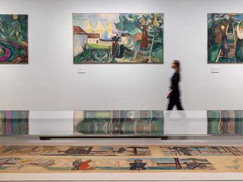 In the foreground is a glass display case containing sketches. In the background, a woman walks along a white wall while looking at the paintings hanging there. Photograph.