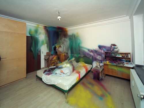 Et rom med en seng midt i og diverse møbler på hver side av sengen. Sengen, deler av rommet og møblene er spraymalet i ulike farger. Fotografi