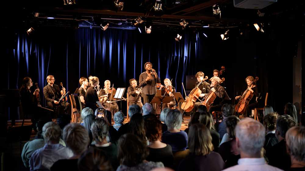 Foto av bandmedlemmer i konsert på scenen med publikum i forgrunnen.