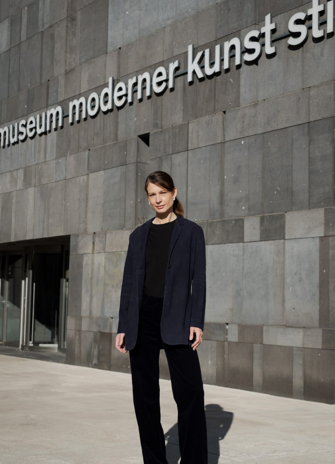 Fatima Hellberg stands outside the Museum moderner Kunst Stiftung Ludwig in Vienna. She is shown full-length, wearing black trousers and a sweater with a dark blue blazer. Photograph.