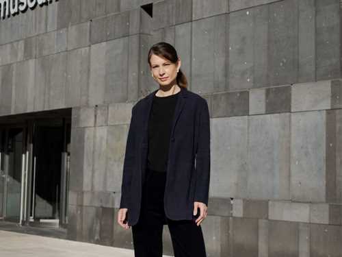 Fatima Hellberg stands outside the Museum moderner Kunst Stiftung Ludwig in Vienna. She is shown full-length, wearing black trousers and a sweater with a dark blue blazer. Photograph.