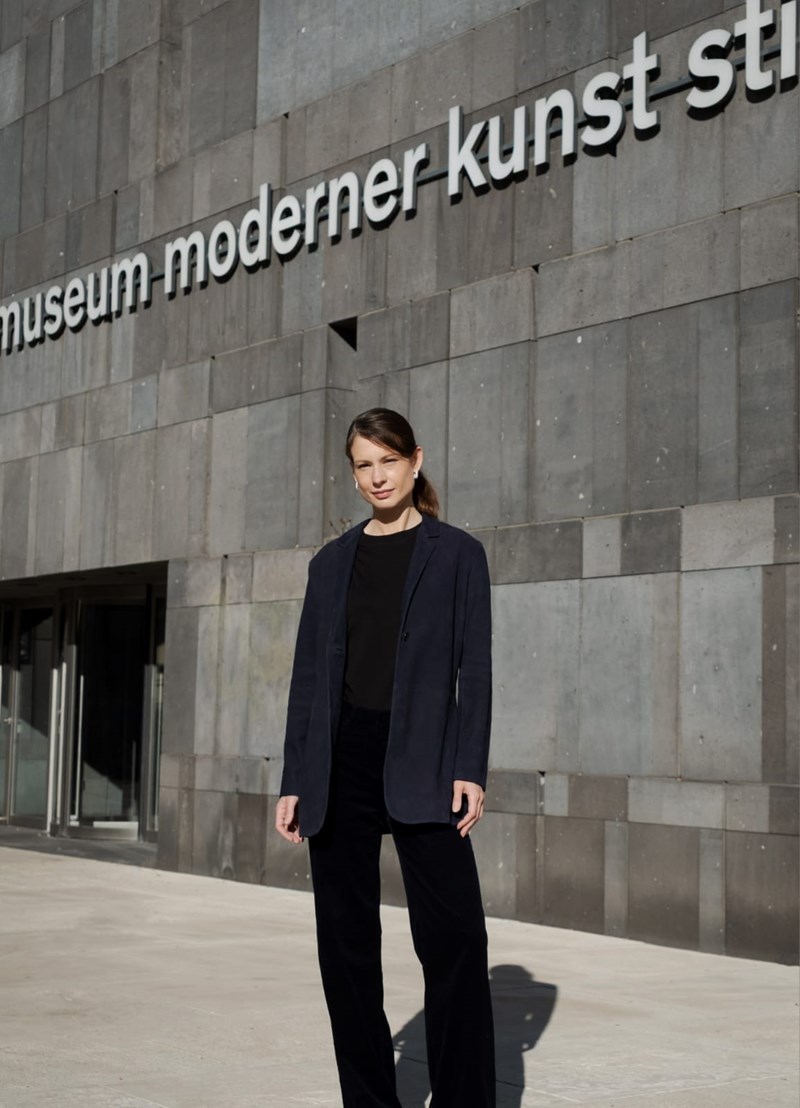 Fatima Hellberg står utenfor Museum moderner Kunst Stiftung Ludwig i Wien. Hun er kledd i svart bukse og genser og har en mørkeblå blazer. Hun er i helfigur. Fotografi
