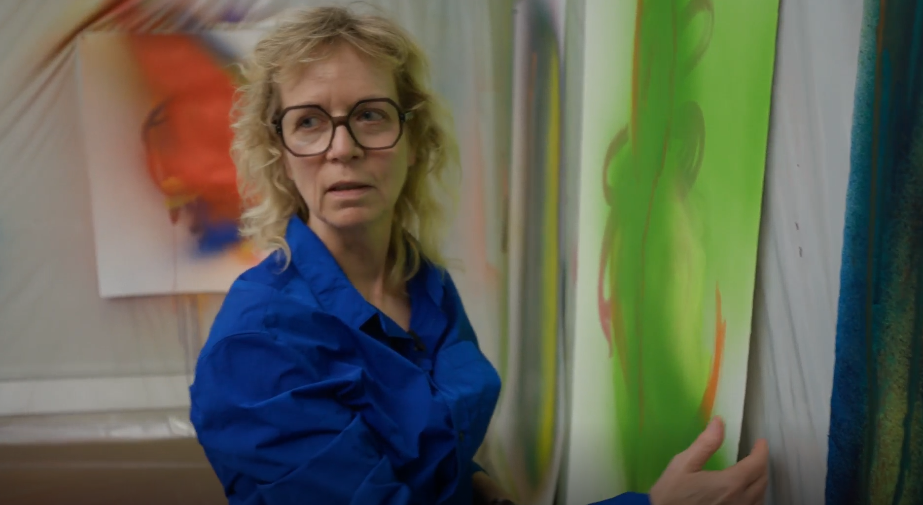 Portrait of a blond woman with big glasses and a blue shirt holding a green painting 