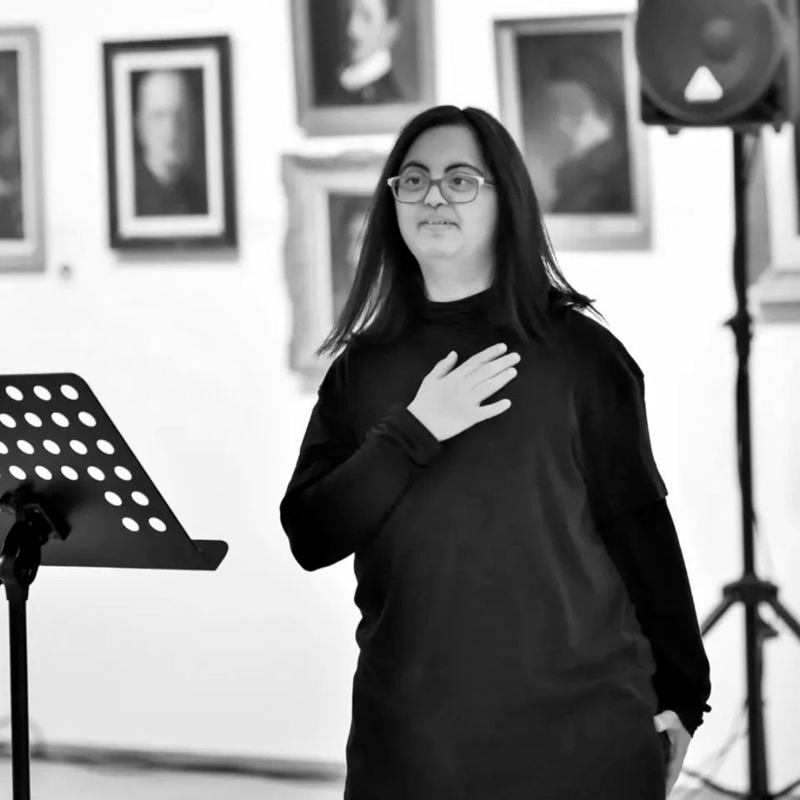 A black-and-white photo of a woman with glasses and long hair, dressed in black, holding a hand to her chest and looking toward the audience. A gallery wall with small artworks is behind her.