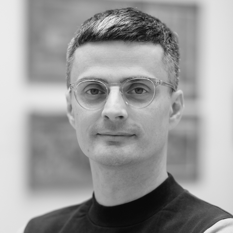 A black-and-white portrait of a man with short hair and round glasses, facing the camera with a calm expression. He wears a two-tone top. Blurred artworks are visible in the background.
