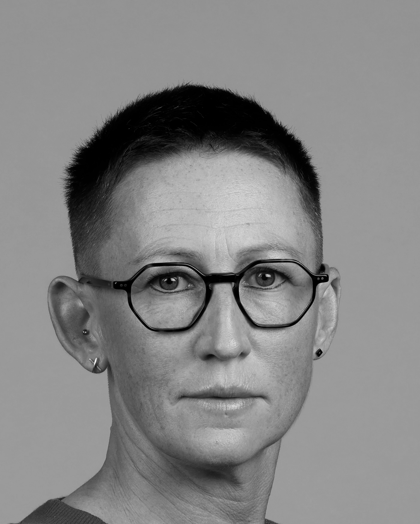 Black and white photo of a person with round glasses, short hair and pierced ears.