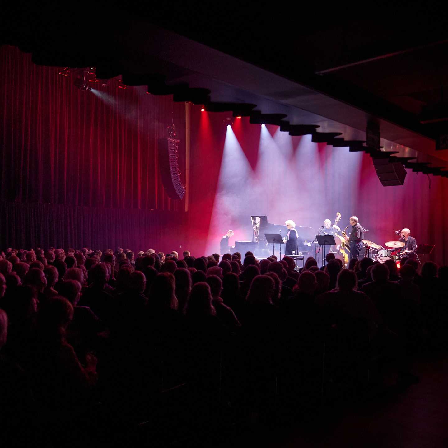 En fullsatt festsal omgitt av mørkt rosa lys, og spotlys mot scenen, hvor en gruppe jazzmusikere skimtes.