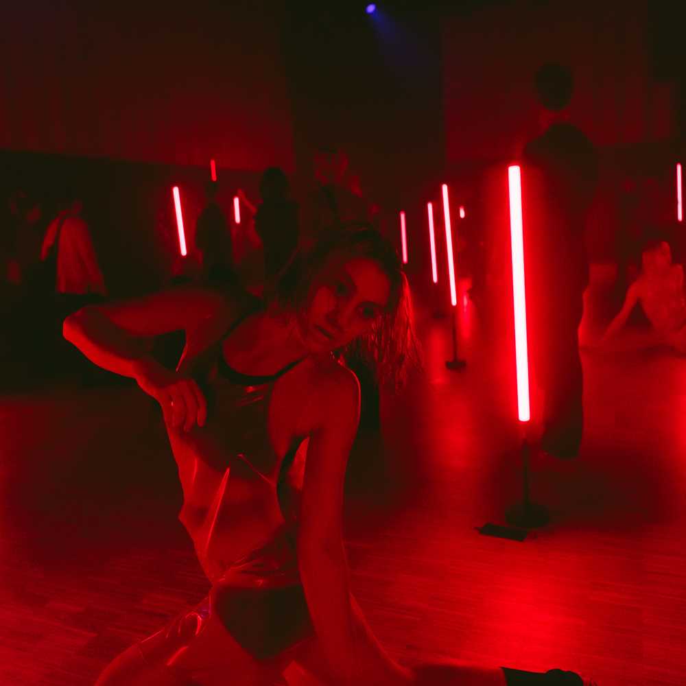 People wearing transparent plastic clothing move around a dark room illuminated by red fluorescent lights.