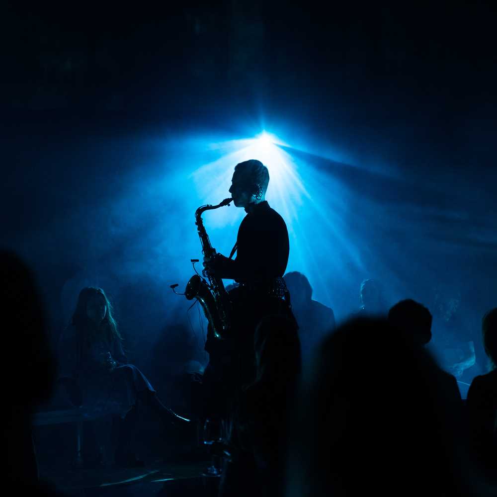 Saxophonist Bendik Giske stands and plays in the middle of a crowd seated on the floor, listening. The room is dark, illuminated by blue light. Photograph.