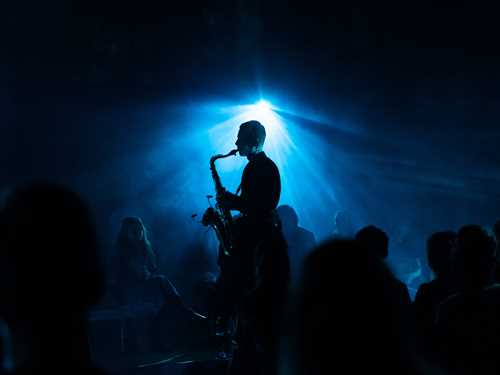 Saksofonist Bendik Giske står og spiller midt i en folkemengde som sitter på gulvet og hører på. Rommet er mørkt og lyst opp av blått lys. Fotografi.