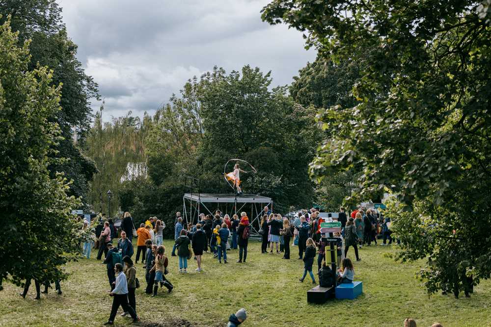 Midt i den grønne og frodige Tøyenparken er det bygget opp en høy scene der en mann danser med flagrenden bånd. En menneskemengde står på gressbakken og ser opp på ham. Fotografi