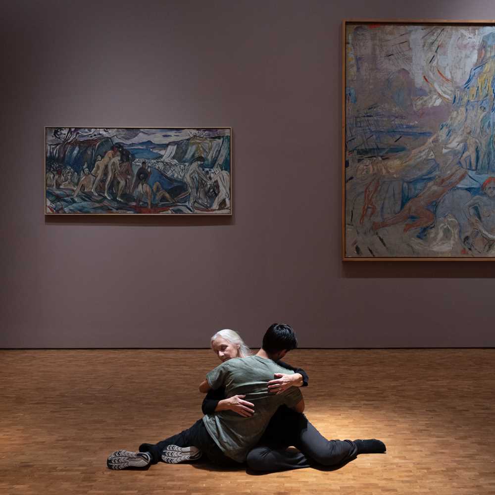 Two people sit on the floor in an embrace. In the background is Edvard Munch's painting The Human Mountain. Photograph.