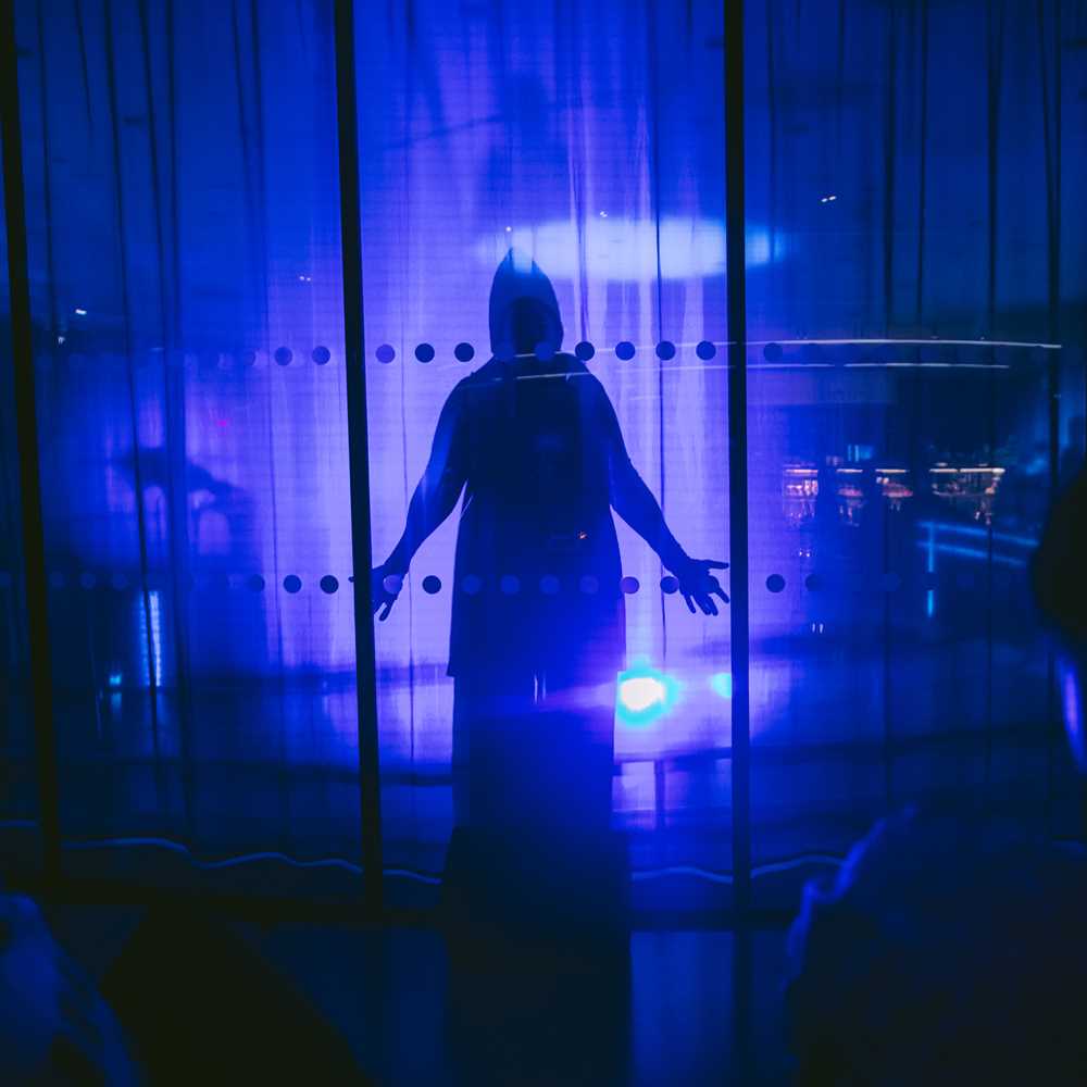 A cloaked figure stands behind a glass wall in a room illuminated by blue light. Photograph.