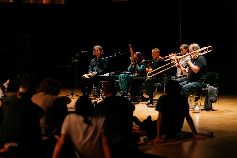 Five people with different instruments sit on individual chairs, performing in front of an audience seated on blankets on the floor. Photograph.