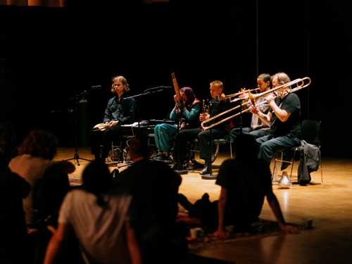 Fem mennesker med ulike instrumenter sitter på hver sin stol og spiller foran et publikum som sitter på tepper på gulvet. Fotografi