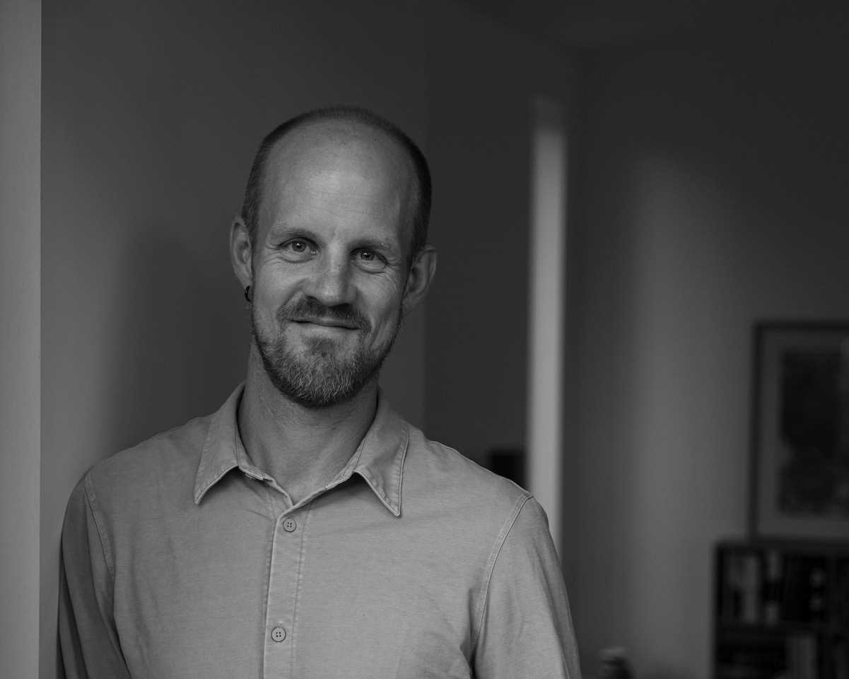 Portrett i sort hvitt av smilende mann med kort hår, lyst skjorte og lett skjegg. 