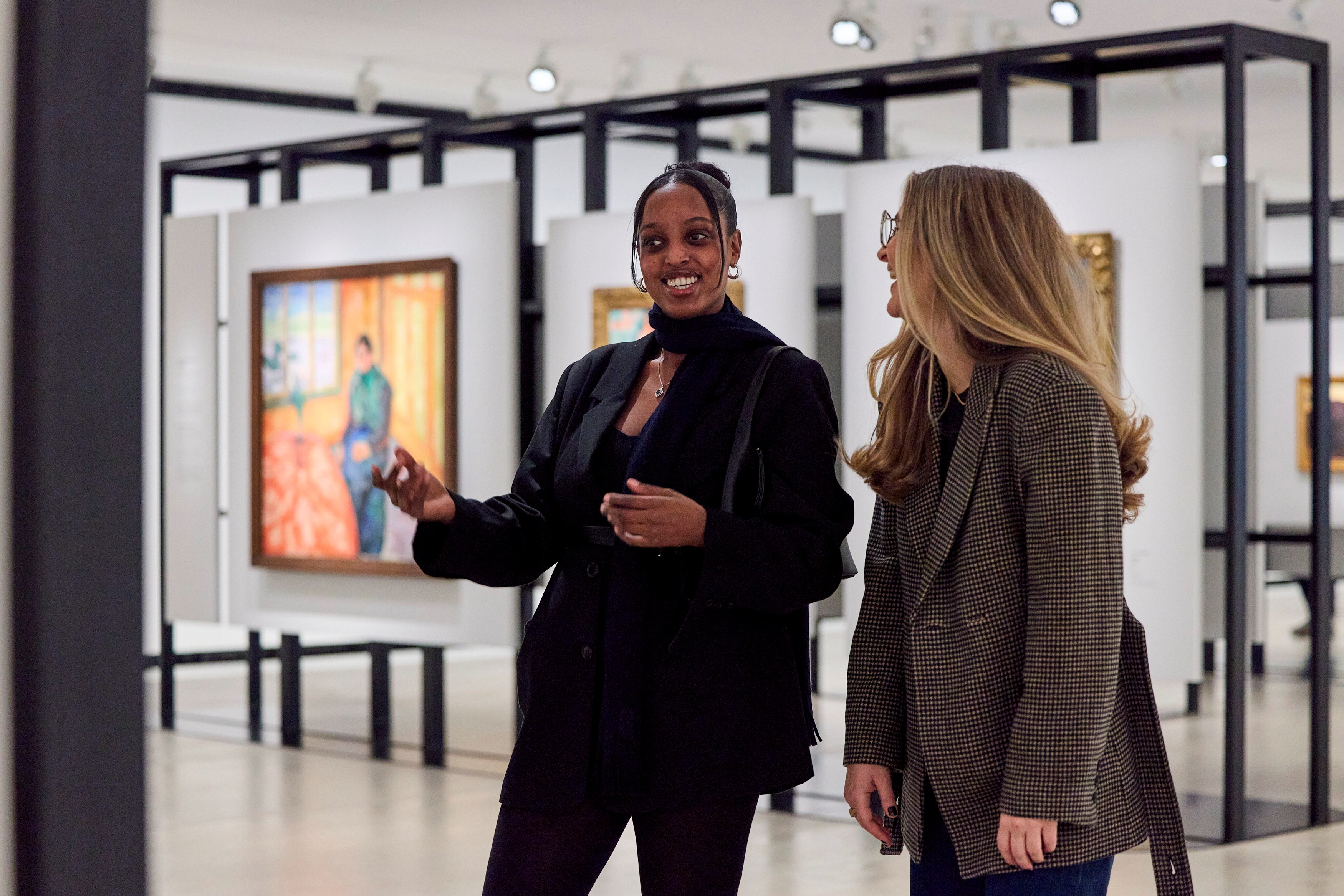 To venner står i et galleri og snakker sammen og ler mens de ser på malerier. Fotografi