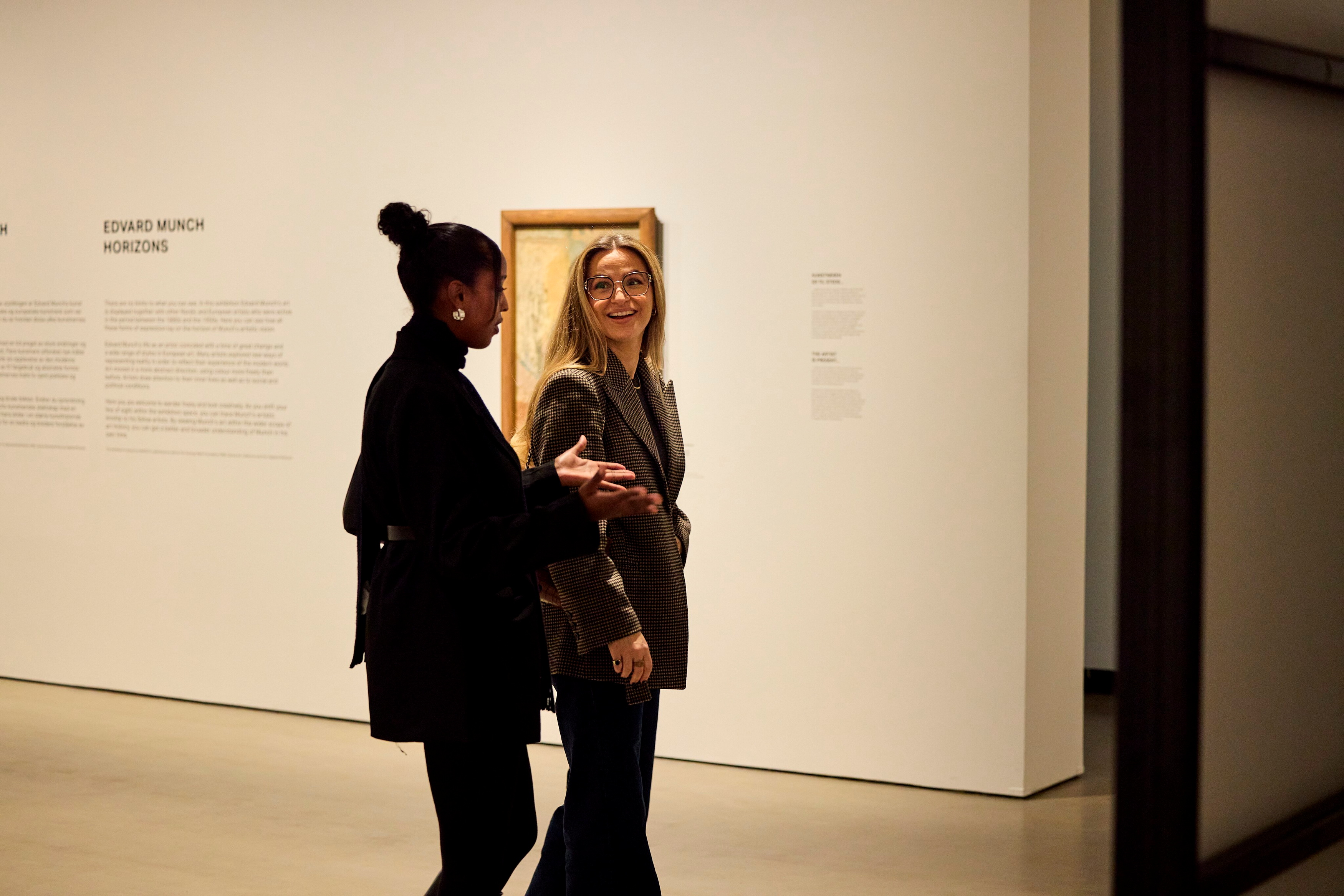 To venner vandrer gjennom en utstillingssal mens de ser på hverandre, har en samtale og gestikulerer og smiler. Fotografi