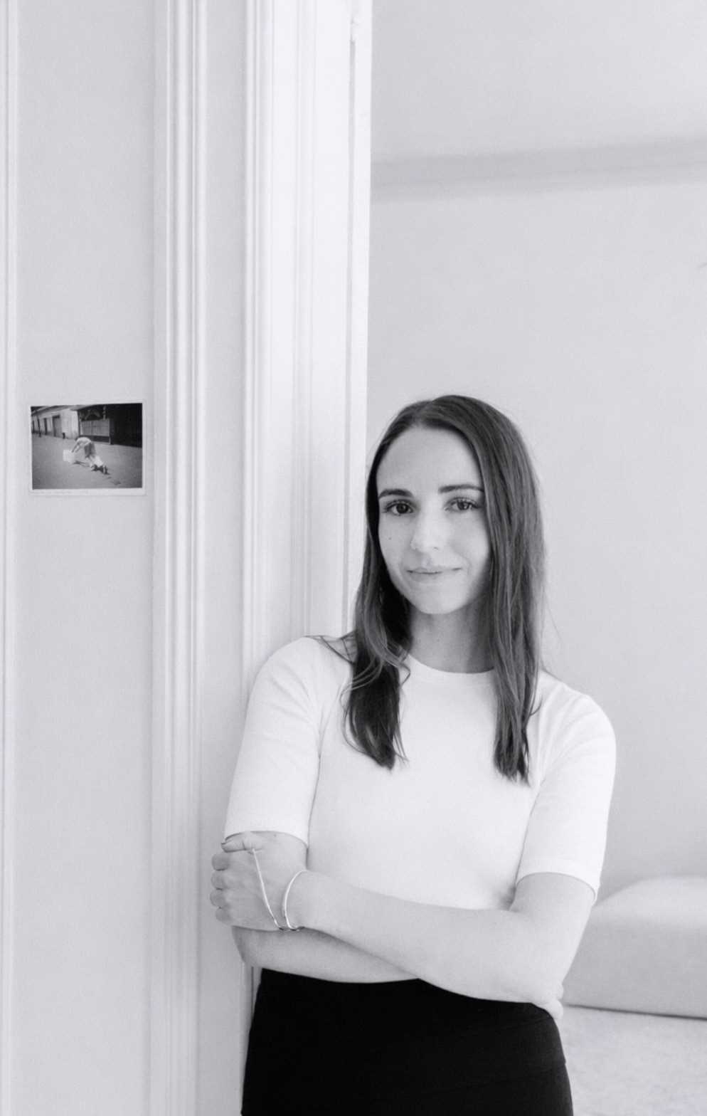 Portrett i sort hvitt av kvinne med brunt langt hår, stående inntil en hvit vegg. 